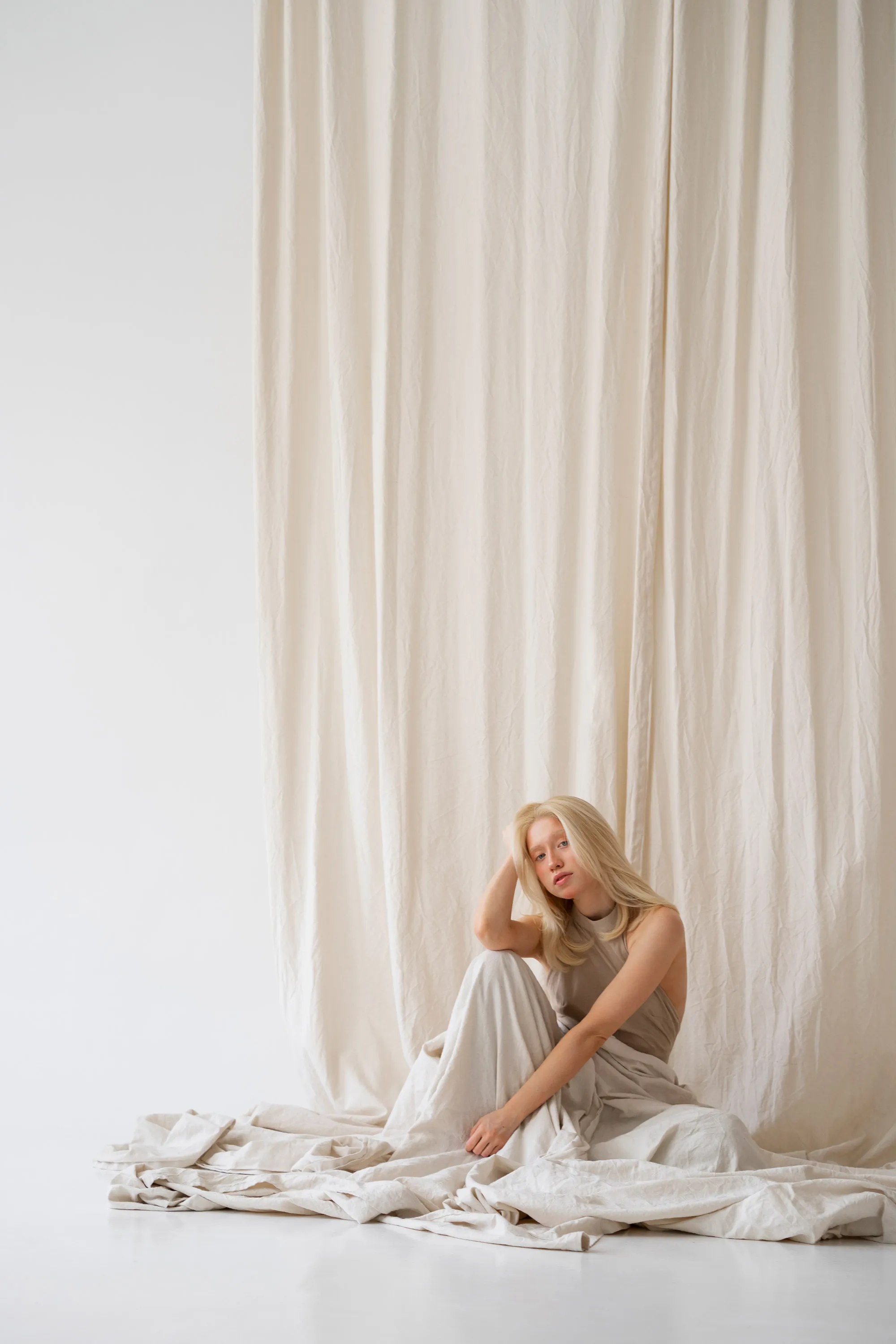 A model with the soft and natural aesthetic of the Marrakech Oil skincare brand, seated amongst flowing neutral-toned fabrics.